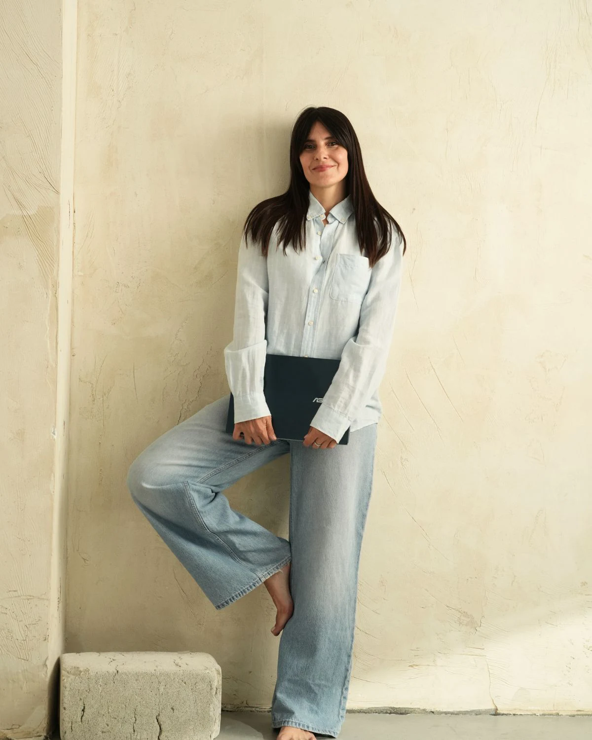 marija sotiji naslonjena na zid, noga je jedna podignuta i naslonjena na drugu, dok u ruci drzi laptop uz blagi osmeh