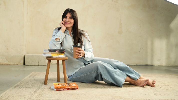 marija maika sedi na pod i drzi kafu u ruci, okruzena knjigama